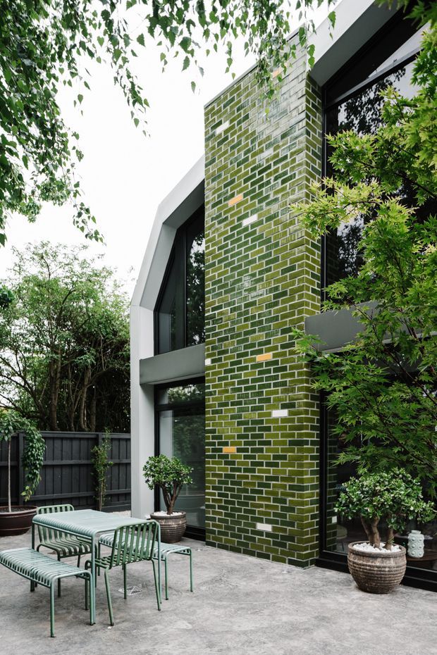 A contemporary courtyard terrace beside a modern two-storey home. The facade features a tall green and white glazed brick column, dark-framed floor-to-ceiling glazing, and smooth light-grey structural surrounds. A green metal outdoor dining set sits on a concrete-paved terrace, surrounded by mature planting and a dark boundary fence.
