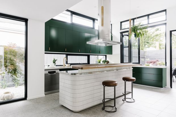 Bright contemporary kitchen with a large curved island clad in small white tiles, dark green flat-panel cabinetry, pale stone-look benchtops, black-framed glazing, and two brown leather stools. The room opens to a planted outdoor area through extensive windows and doors, with pendant lighting and stainless-steel appliances.