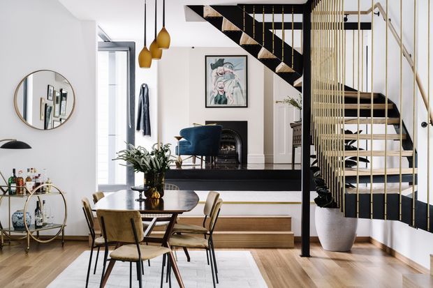 A bright, refined open-plan interior centered on a dining area, with a round dark timber dining table and upholstered timber chairs in the foreground. A contemporary black-and-white staircase with open risers, slim gold-toned vertical balustrade elements, and a black structural frame dominates the right side. The space includes light timber flooring, white walls, a linear timber console, sculptural pendant lights, a blue armchair, artwork, a bar cart, and indoor planting.