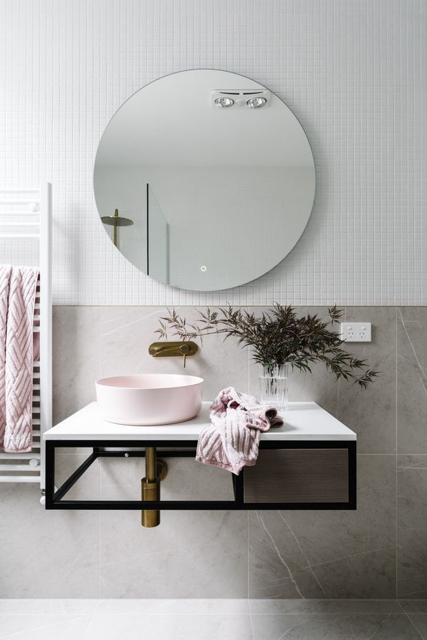 A refined contemporary bathroom vanity area featuring a wall-mounted dark vanity with a white countertop, pale pink vessel basin, brass wall-mounted tapware and exposed brass plumbing. A large round illuminated mirror hangs above, set against small white grid tiles. Large pale grey stone-look wall tiles continue below the tile line. A white heated towel rail with a soft pink towel is visible at left, adding a warm accent.
