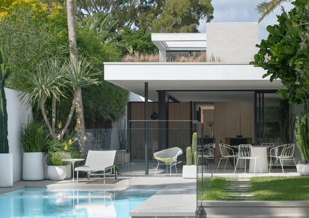 Contemporary flat-roofed residence viewed from the landscaped pool area, with expansive floor-to-ceiling glazing, a deep white roof overhang, black-framed glass walls, and a white brick upper volume. The outdoor space includes a swimming pool, pale paved terrace, clipped lawn, glass balustrade, and tropical planting. Through the glazing, a timber-lined interior with dining furniture is visible.