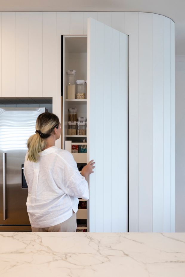 A bright contemporary kitchen with full-height white vertically panelled cabinetry and a tall pull-out pantry open to reveal organised shelves of jars and dry goods. A stainless-steel refrigerator is partly visible at left, while a large white marble-look island benchtop with grey veining fills the foreground. A person is opening the pantry door.