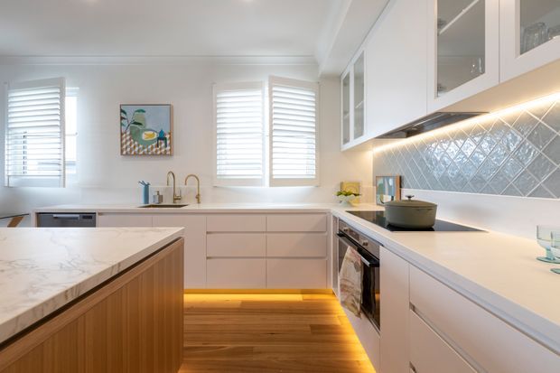 Bright, refined contemporary kitchen with white cabinetry, glass-front upper cupboards, a marble-look island benchtop, and a blue-grey diamond-pattern tiled splashback. Shaker-style cabinetry, under-cabinet lighting, timber flooring, plantation shutters, brass tapware, and warm toe-kick lighting create a welcoming, coastal-leaning interior.