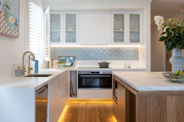 Bright, refined kitchen photographed from the entry side, featuring white perimeter cabinetry with glass-front upper doors, a pale marble-look island benchtop, timber island cabinetry, integrated appliances, a stainless-steel sink with brushed brass tapware, and a blue-grey patterned tile splashback. Warm under-cabinet and toe-kick lighting highlights the timber flooring. A window with white plantation shutters provides natural light, while a floral arrangement and artwork add soft decorative colour.
