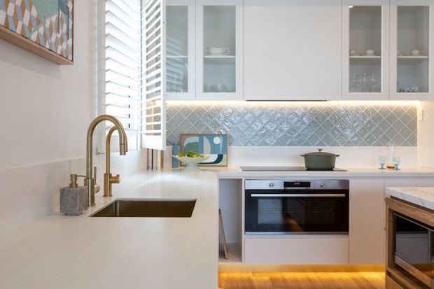 Bright contemporary kitchen with white cabinetry, integrated glass-front display cupboards, a pale stone benchtop, undermount sink and brass gooseneck tap. A decorative blue-grey patterned tile splashback runs beneath the upper cabinets, which feature warm under-cabinet lighting. The kitchen includes a built-in oven, cooktop and timber-accented island or peninsula, with plantation shutters and colourful geometric artwork adding visual interest.