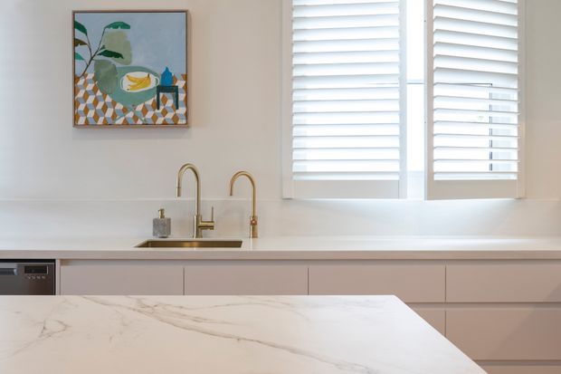 Bright contemporary kitchen detail featuring pale flat-panel cabinetry, a white veined stone benchtop in the foreground, a white tiled or painted backsplash, and brass-toned gooseneck and secondary taps. Plantation shutters cover the windows, while a colourful framed botanical artwork adds a small accent above the sink.