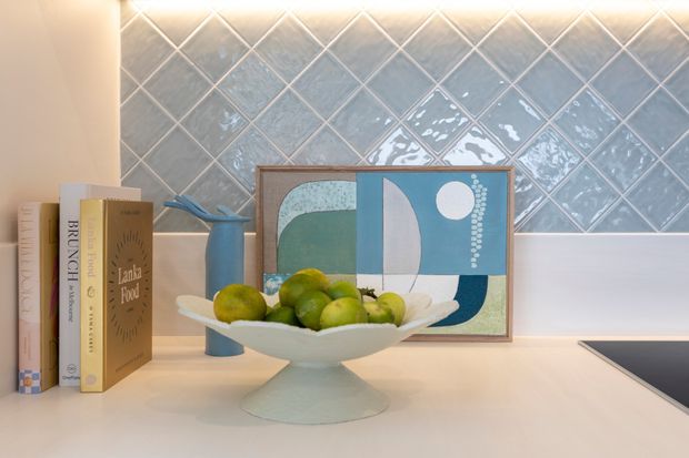 Close-up of a styled kitchen benchtop with a pale solid surface, a raised white fruit bowl, cookbooks, a blue vase, and framed abstract artwork. The backdrop is a glossy blue-grey diamond-pattern tiled splashback with under-cabinet lighting; a black cooktop edge is visible at right.