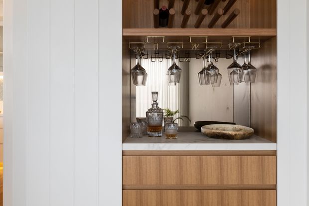 Close-up of a built-in timber drinks cabinet or home bar. The open upper section has integrated wine-bottle storage and hanging racks for stemware, with decanters and glasses displayed on a grey benchtop. Warm timber drawers sit below, framed by tall white wall panelling.
