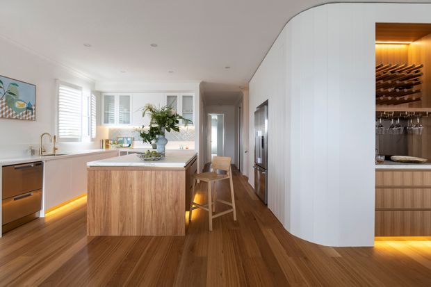 Bright, refined contemporary kitchen with warm timber flooring and island cabinetry, white integrated joinery, a pale benchtop, brass-toned tapware, and under-cabinet lighting. A curved white wall conceals a prominently illuminated built-in wine cellar with bottle storage and stemware racks. The kitchen includes a central island, integrated appliances, bar seating, timber floors, and ample natural light from windows and shutters.