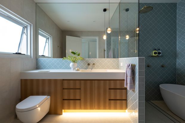 Contemporary bathroom featuring a freestanding bathtub behind a glazed shower screen, a wall-mounted timber vanity with integrated drawers and warm under-cabinet lighting, pale geometric wall tiles, large mirrors, and high horizontal awning windows. Pendant lights and brass-toned fixtures add warmth to the clean, refined scheme.