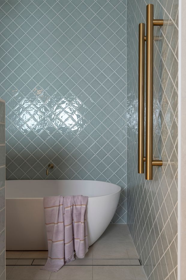 A compact, refined bathroom featuring a freestanding white oval bathtub set against pale blue-grey glossy square tiles installed diagonally. Large brushed brass/gold wall-mounted heated towel rails or fixtures occupy the right wall, while large light-grey floor tiles and soft lavender towels add subtle contrast.