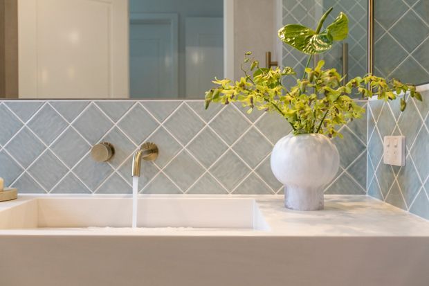 Close-up view of a refined bathroom vanity with a rectangular white basin, brushed brass wall-mounted tapware, pale stone benchtop, and grey-blue diamond-pattern tiled splashback. A large mirror reflects the tiled wall and adjacent cabinetry, while a sculptural white vase with leafy branches adds a natural decorative element.