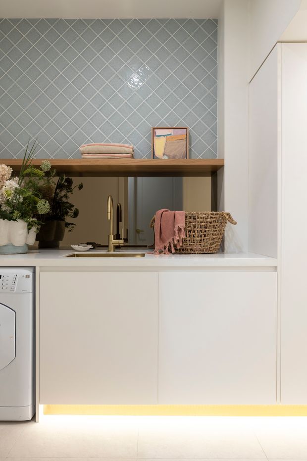 A close-up view of a refined contemporary kitchen or compact utility kitchen, featuring white flat-panel cabinetry, a pale stone-look benchtop, integrated under-cabinet lighting, a stainless sink with brushed brass/gold tapware, and a warm timber open shelf. The backsplash is clad in diagonally laid grey-blue ceramic tiles with light grout. Decorative plants, folded textiles, framed artwork, and a woven basket add warmth, while a partially visible front-loading appliance sits at left.