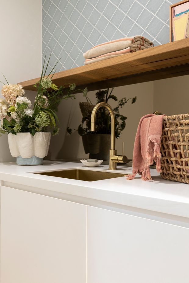 Close-up of a refined bathroom vanity with white fixed cabinetry and a white benchtop, inset rectangular basin, and brushed brass gooseneck tapware. A large frameless mirror reflects greenery, while an open timber shelf above holds folded towels and artwork. Decorative flowers, woven storage, and a blush pink towel add warmth and texture. The wall behind the shelf is finished with pale grey-blue diagonal grid tiles.