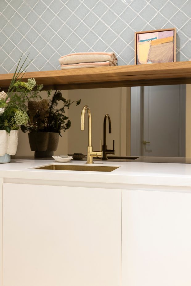 Close-up view of a refined kitchen sink area with white integrated cabinetry and a white benchtop, dual warm metallic and dark taps, and a rectangular undermount sink. A large mirror or reflective splashback sits behind the sink, reflecting the cabinetry and doorway. Above it is a substantial open timber shelf displaying folded towels, framed artwork, and potted greenery. The upper wall is clad in pale grey diamond-patterned tiles, creating a warm, contemporary decorative backdrop.