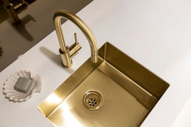 Close-up product-style view of a polished brass or gold-toned undermount kitchen sink with a matching high-arched gooseneck mixer tap, set into a clean white benchtop. A small decorative soap dish sits beside the sink, and the reflective surface creates a warm, luxurious appearance.