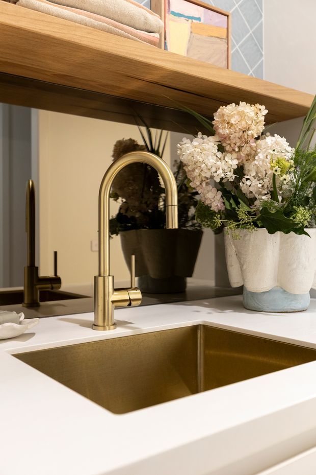 Close-up of a refined kitchen sink area featuring a large undermount metallic sink, warm brushed-gold gooseneck mixer tap, matching secondary tap, and white engineered benchtop. A timber open shelf spans the upper frame, with folded textiles, framed artwork, a black planter, and a large floral arrangement adding warmth. A mirror or reflective splashback behind the sink reflects the planter and room. The palette is warm, neutral, and softly styled.
