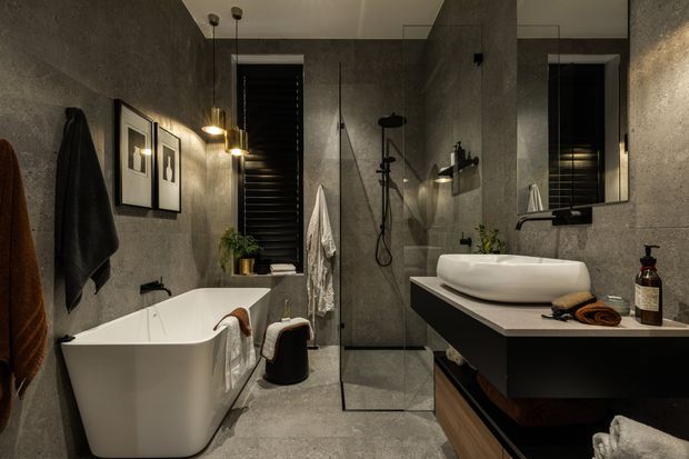 A moody contemporary bathroom with dark textured grey walls and matching floor, a freestanding white bathtub, frameless glass shower enclosure, wall-mounted black fixtures, and a floating dark vanity with a white vessel basin. Warm pendant lighting, timber accents, towels, framed artwork, and small plants add contrast and texture.