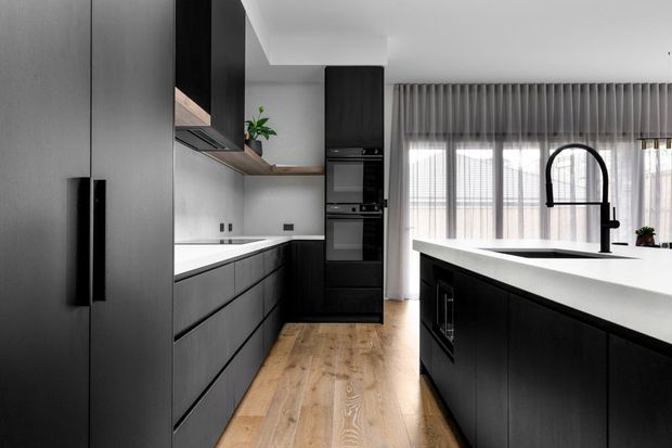 Contemporary galley-style kitchen with dark charcoal cabinetry, a large white island, integrated black appliances, timber flooring, and full-height sheer curtains over a bright window wall. A striking arched black gooseneck tap and simple indoor greenery add detail to the clean, high-contrast space.