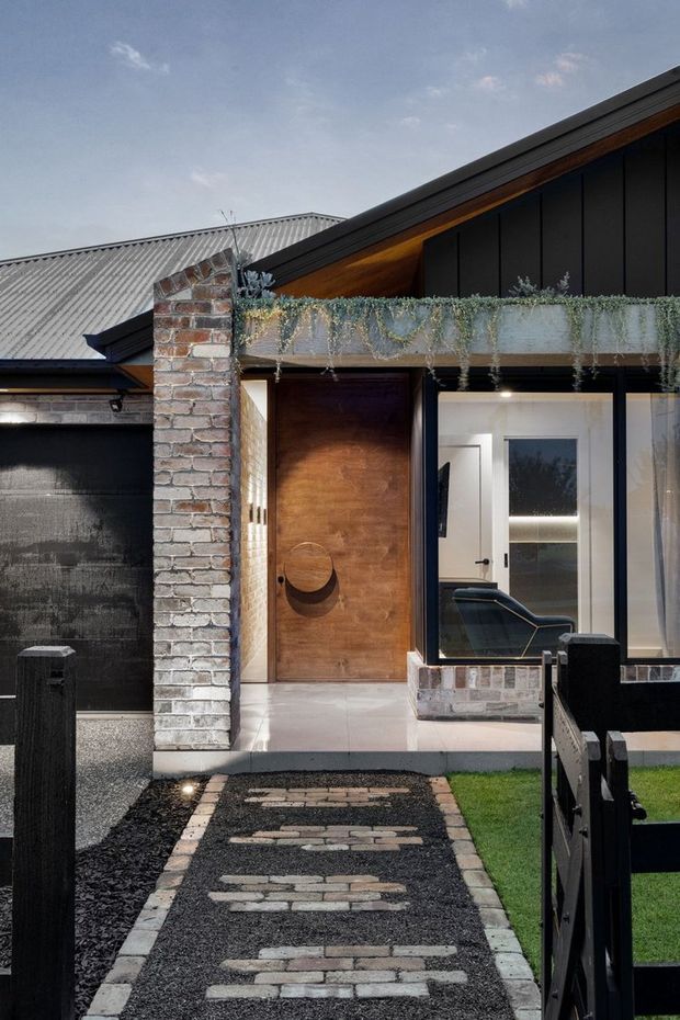 Exterior view of a contemporary residential entry, featuring a dark vertical-clad facade, exposed brickwork, a warm timber pivot door, black-framed glazing, and a planted concrete canopy. A gravel-and-brick path leads through a small front garden to the entrance.