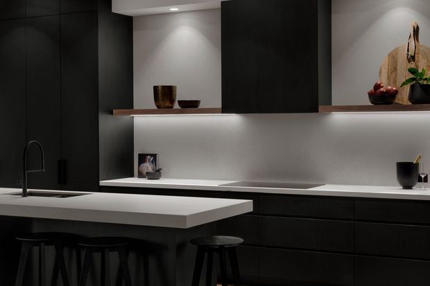 A dark, refined contemporary kitchen featuring matte-black full-height cabinetry, a pale stone island and benchtop, integrated appliances, a black undermount sink and tap, and softly illuminated open display shelving with minimal decorative objects. The space has a restrained, moody palette and streamlined built-in joinery.