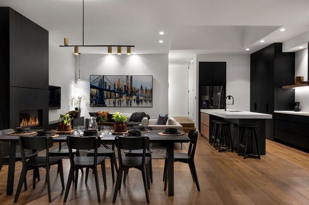 Open-plan contemporary kitchen, dining, and living space with dark built-in cabinetry, a white island benchtop, timber flooring, black dining furniture, recessed lighting, and a linear fireplace feature. A large artwork and lounge seating are visible beyond the dining area.