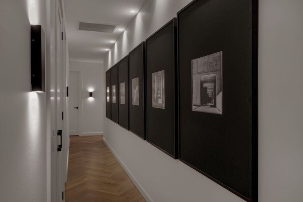 A narrow, elegantly styled residential hallway with white walls, recessed ceiling lights, and a warm herringbone timber floor. A long series of large black-framed artworks lines the right wall, while white paneled doors and minimal wall sconces define the corridor. The overall composition is monochromatic, refined, and gallery-like.