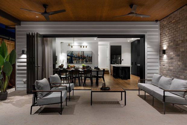 A covered outdoor living area opens directly into a contemporary dining and kitchen space through wide glazed sliding doors. The outdoor zone has a timber-lined ceiling with ceiling fans, pale concrete flooring, light upholstered lounge seating, and a black metal coffee table. A white horizontal-clad wall and exposed brick wall frame the opening, while the interior features a dining table, kitchen island, timber flooring, dark cabinetry, and modern pendant lighting.