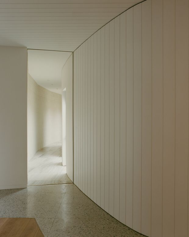 A minimalist interior hallway with a gently curving wall and ceiling clad in narrow vertical pale timber or painted panels. The corridor opens toward a softly lit passage with curved walls and light flooring. The foreground floor is polished terrazzo, with a small section of warm timber flooring visible at the lower left. The palette is restrained in off-white, cream, and light grey, creating a calm, sculptural atmosphere.