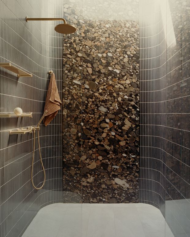 A compact contemporary walk-in shower with dark grey vertically gridded wall tiles, a central full-height pebble mosaic feature wall, and pale large-format floor tiles. Brushed brass shower fittings, a slim overhead rain shower, wall-mounted hand shower, and simple timber-toned accessories add warmth. The curved tile layout and softly lit natural-material feature create an earthy, refined atmosphere.
