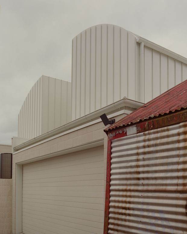 Exterior architectural detail showing a pale industrial or commercial building with a large sectional garage door, light rendered masonry walls, and tall curved corrugated metal roof forms. A weathered corrugated metal structure with a rust-red roof occupies the foreground, creating a contrast between new and aged industrial fabric. The image is softly lit under an overcast sky.
