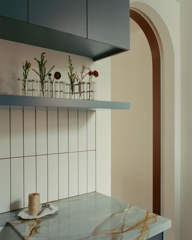 A close-up view of a refined kitchen featuring a pale veined stone benchtop, vertically tiled splashback, dark blue-grey overhead cabinetry, and a matching open shelf displaying glass flower vases. A sculptural arched doorway with a warm timber-toned inner reveal is visible to the right. The palette is muted and warm, combining cool cabinetry with cream walls and dramatic natural stone veining.