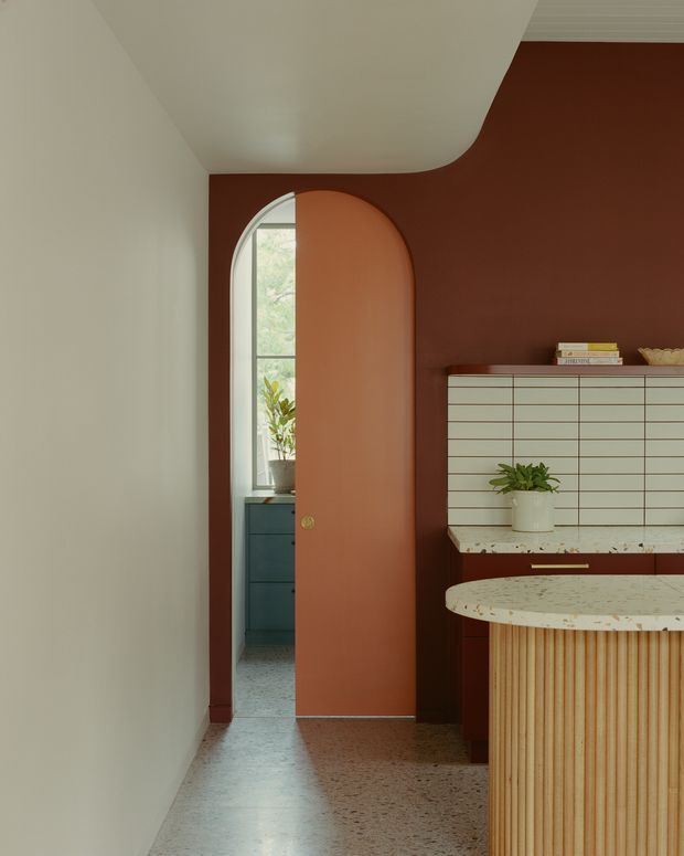A narrow, refined interior passage leading to a rounded-top terracotta door. The space features deep rust-red walls, curved white ceiling detailing, a terrazzo floor, and a built-in kitchenette or bar area with pale horizontal tile cladding, a speckled stone benchtop, and fluted timber cabinetry. A tall arched window and blue cabinetry are visible beyond the doorway. The composition emphasizes soft curves, warm earthy colours, and integrated joinery.