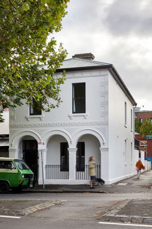 Street-level exterior view of a narrow two-storey white masonry building with ornate decorative mouldings, repeated arched verandah openings, black wrought-iron fencing, tall sash-style windows, and a corrugated metal hipped roof with chimney. The building occupies a leafy urban corner site beside paved streets and neighbouring buildings.