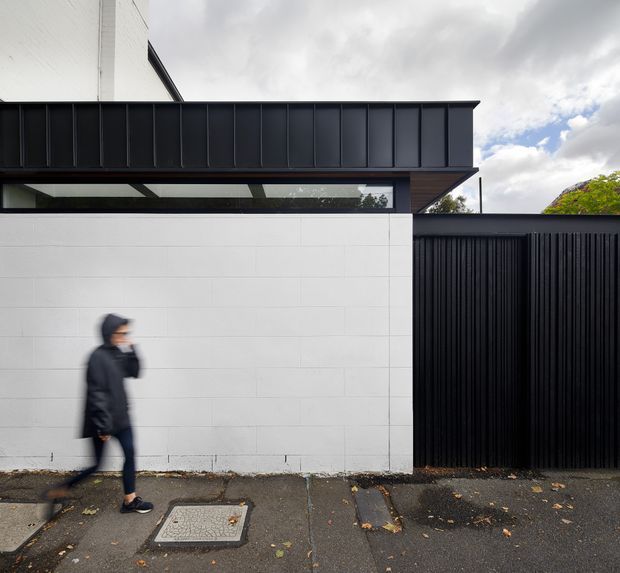 Street-level exterior view of a contemporary building with a bold black metal parapet and projecting flat roof, a long horizontal glazed clerestory, white masonry wall panels, and a tall black vertical-slat gate. A blurred pedestrian and paved footpath appear in the foreground beneath an overcast sky.