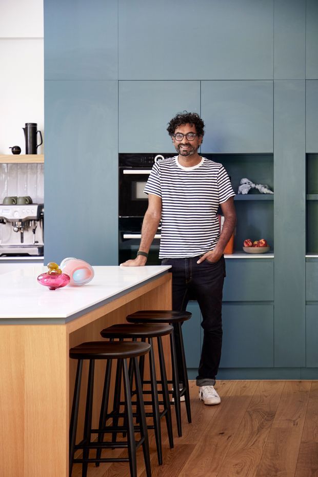 A contemporary kitchen featuring full-height muted blue-green cabinetry, integrated black appliances, open shelving, a large white island with a timber base, and black counter stools. Warm timber flooring and a person standing at the island add scale and a lived-in feel.