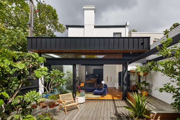 Exterior view of a modern residential extension opening onto a timber deck and lush planted courtyard. The dark metal-clad upper balcony and structural framing contrast with the white rendered masonry house, while large glazed doors reveal a colourful living room inside.