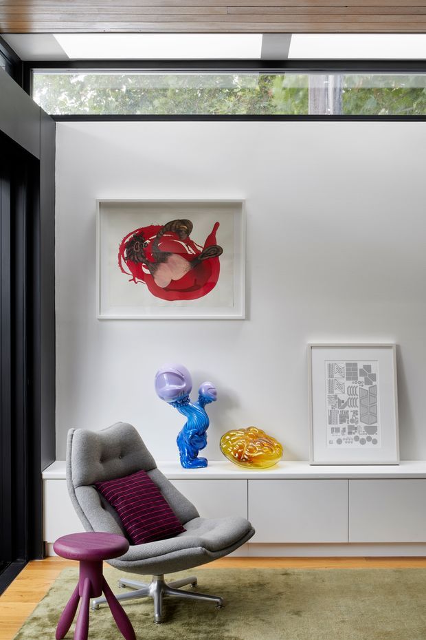 A refined contemporary interior vignette featuring a grey upholstered lounge chair with a magenta cushion and matching sculptural side table. White built-in cabinetry and display shelving line the wall, styled with colourful glass objects and framed artwork. Black-framed clerestory glazing and a timber-lined ceiling introduce strong architectural contrast, while a muted green rug grounds the seating area.
