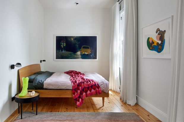 A bright, compact bedroom with a low timber bed and rounded timber headboard, white walls, and honey-toned timber floorboards. Floor-to-ceiling sheer curtains line the tall window, while black wall-mounted reading lights, framed artwork, a small black side table, layered bedding, and a patterned throw add character. The room has a clean contemporary feel with warm natural timber and colourful textile accents.