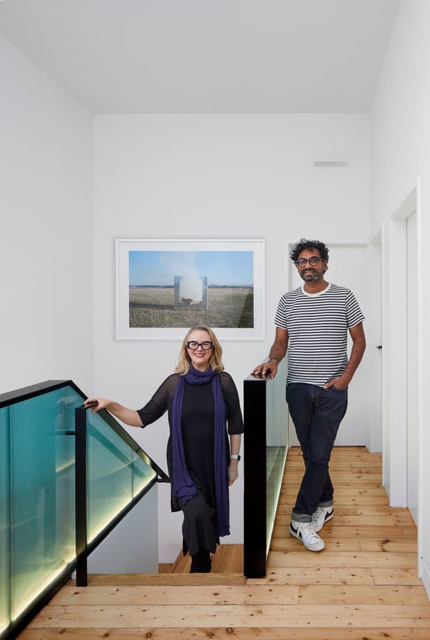 A bright, minimalist interior landing at the top of a staircase, featuring white walls and ceiling, pale timber flooring, and a contemporary glass balustrade with dark framing. Two people stand on the landing beside the stair opening. A framed landscape artwork hangs on the wall, and doorways lead to adjacent rooms.