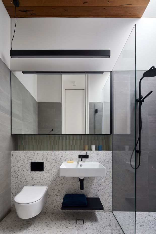 Contemporary bathroom with a walk-in glass shower, wall-mounted white toilet and basin, large mirrored cabinet, terrazzo wall and floor surfaces, grey tiled shower enclosure, dark plumbing fixtures, and a timber-lined ceiling with a suspended black linear light.