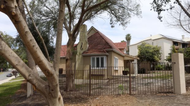 Street-facing view of a traditional single-storey house behind a metal fence, with cream rendered walls, terracotta tiled pitched roofs, timber-look louvred shutters, and a small front garden. Mature trees partially obscure the facade, while neighbouring homes are visible in the background.