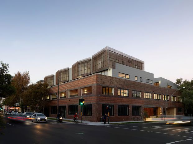Twilight street-level view of a multi-storey urban building with a strong brick podium and contemporary upper levels. The lower floors feature dark brick masonry, large rectangular windows and recessed lighting, while the upper storeys combine pale rendered or metal-clad walls with projecting volumes enclosed by vertical timber-toned screening. Traffic and street markings are visible in the foreground, with trees lining the surrounding urban streetscape.
