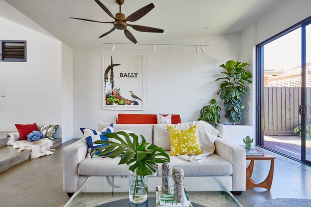 Bright contemporary living room with a white-painted brick feature wall, polished concrete flooring, a large sliding glass door opening to a small courtyard, and a white sofa decorated with colourful cushions. A sculptural wood ceiling fan, indoor plants, framed artwork, and a low timber coffee table create a relaxed, tropical-inspired interior.