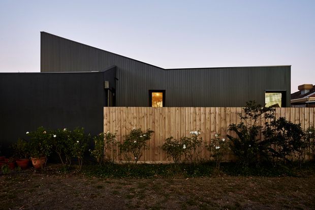 Exterior view of a compact contemporary building with dark vertical metal cladding and a sharply sloping roofline. A tall natural timber privacy fence and planted garden occupy the foreground, with a few illuminated windows visible in the dark facade.