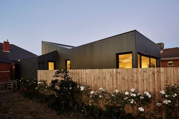 A contemporary single-storey extension or dwelling viewed from a residential backyard at dusk. The building has a sharply angular pitched roof form, dark vertically ribbed metal cladding, and large black-framed windows glowing with warm interior light. A timber paling fence and flowering garden occupy the foreground, with neighbouring brick homes visible on either side.