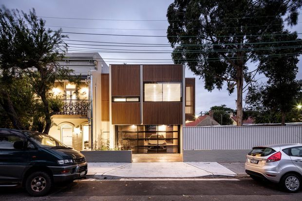 Street-facing view of a contemporary multi-storey urban residence at dusk. The narrow facade combines vertical timber-look screening, large glazed openings, and a dark-framed ground-floor entry or garage-like opening. An ornate traditional neighbouring building is visible to the left, with mature trees, overhead utility wires, a pale boundary fence, sidewalk, and parked vehicles in the streetscape.