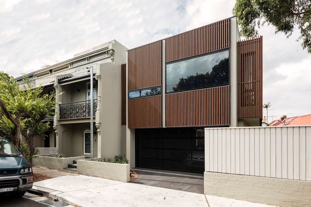 Street-facing view of a contemporary multi-storey residence with a prominent vertical timber batten facade, large dark-framed glazing, an integrated black garage door, and a white rendered heritage-style neighbouring facade with balcony detailing. The home sits along a suburban urban streetscape with a low masonry boundary wall and driveway.