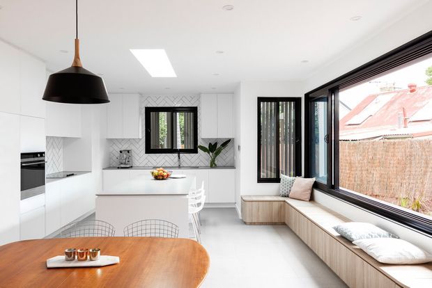 Bright contemporary open-plan kitchen and dining area with white flat-panel cabinetry, a central island, integrated appliances, a tiled geometric splashback, and a large warm timber dining table. Black-framed windows and doors open to a built-in timber window seat with cushions, creating a light-filled connection to the exterior. A black pendant light provides contrast, while a skylight and recessed ceiling lights enhance the airy space.