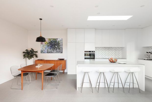 Bright contemporary open-plan kitchen and dining area with a crisp white palette. The space features a large white island with integrated seating, white flat-panel cabinetry, a herringbone-patterned tiled splashback, pale flooring, and a timber dining table with wire-frame chairs. A skylight, recessed ceiling lights, and a black pendant add contrast, while a timber sideboard and indoor plant bring warmth.